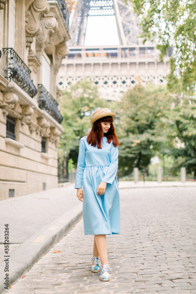 Pretty woman tourist, wearing stylish hat and blue dress, walking on the cozy street with old vintage buildings in old European city Paris, with Eiffel tower on the background
