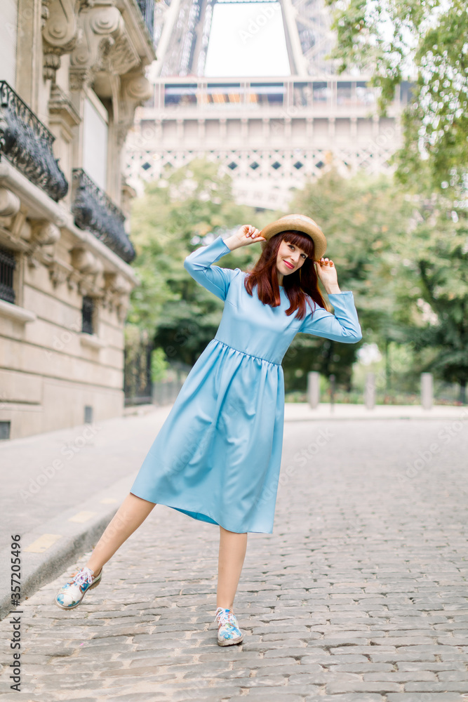 pretty young red haired lady, wearing elegant blue dress and straw hat, walking in Paris, posing to camera and having fun. Eiffel tower behind the woman