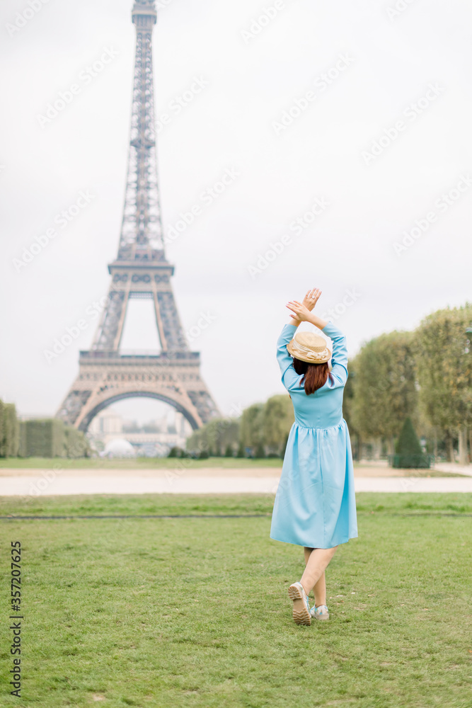 Back view of pretty lady in blue dress and hat, enjoying walk in Paris, near Eiffel tower. Young attractive happy woman with hands up facing the Eiffel Tower in Paris, France.