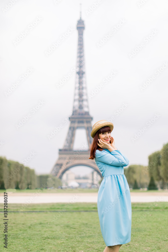 Cheerful smiling woman wearing blue dress and straw hat posing to camera with smile, while standing on green grass in park in front of Eiffel Tower, Paris