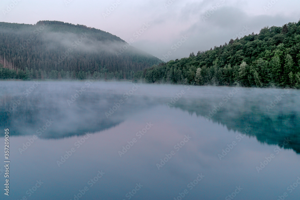 Green looking lake with trees over the early misty morning water in the touristic Belgian Ardennes Green looking lake with trees over the early misty morning water in the touristic Belgian Ardennes