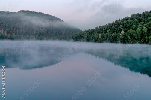 Green looking lake with trees over the early misty morning water in the touristic Belgian Ardennes