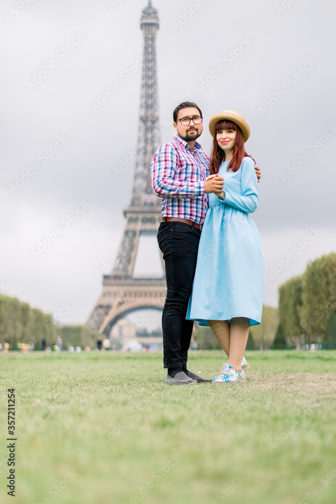 Beautiful young Caucasian couple, pretty red haired woman in blue dress and handsome bearded man in checkered shirt, standing embracing in front of Eiffel tower in Paris. Love in Paris