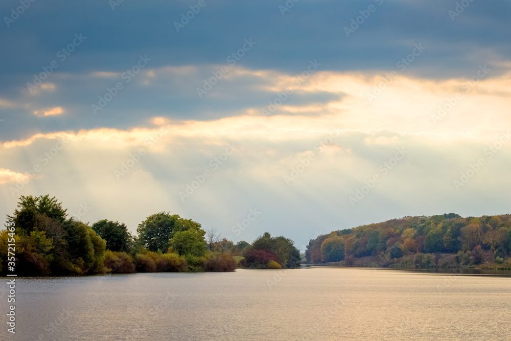 Sunlight penetrates the dark clouds over the river, the river and the forest in the distance with a picturesque sky