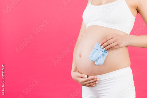 Close up portrait of pregnant woman in white underwear holding blue socks for a baby boy at pink background. Future child expectation concept. Copy space © sosiukin