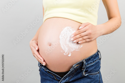 Close up of pregnant woman in unzipped jeans applying moisturizing cream on her belly against stretch marks at colorful background with copy space. Skin care concept © sosiukin