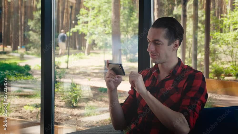 Portrait of handsome young man using his smartphone while sitting on sofa in modern office. Guy in a plaid shirt scrolling phone screen while spending free time. Concept of leisure and tech Portrait of handsome young man using his smartphone while sitting on sofa in modern office. Guy in a plaid shirt scrolling phone screen while spending free time. Concept of leisure and tech