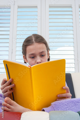 Young girl wearing purple unicorn jumpsuit sitting next to window with a yellow book.