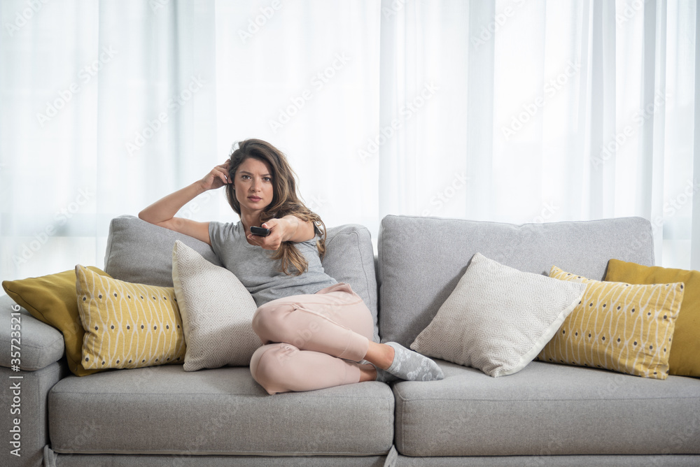 Young beautiful girl sitting on the sofa in apartment changing the tv channels with remote control searching some good television show and program while she having free day from her work Young beautiful girl sitting on the sofa in apartment changing the tv channels with remote control searching some good television show and program while she having free day from her work