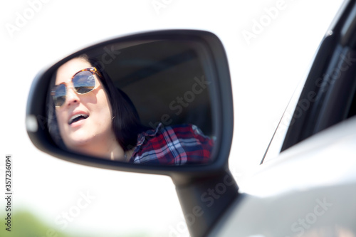 Reflection on a rear view mirror of a young lady having fun driving a car in a sunny summer day.