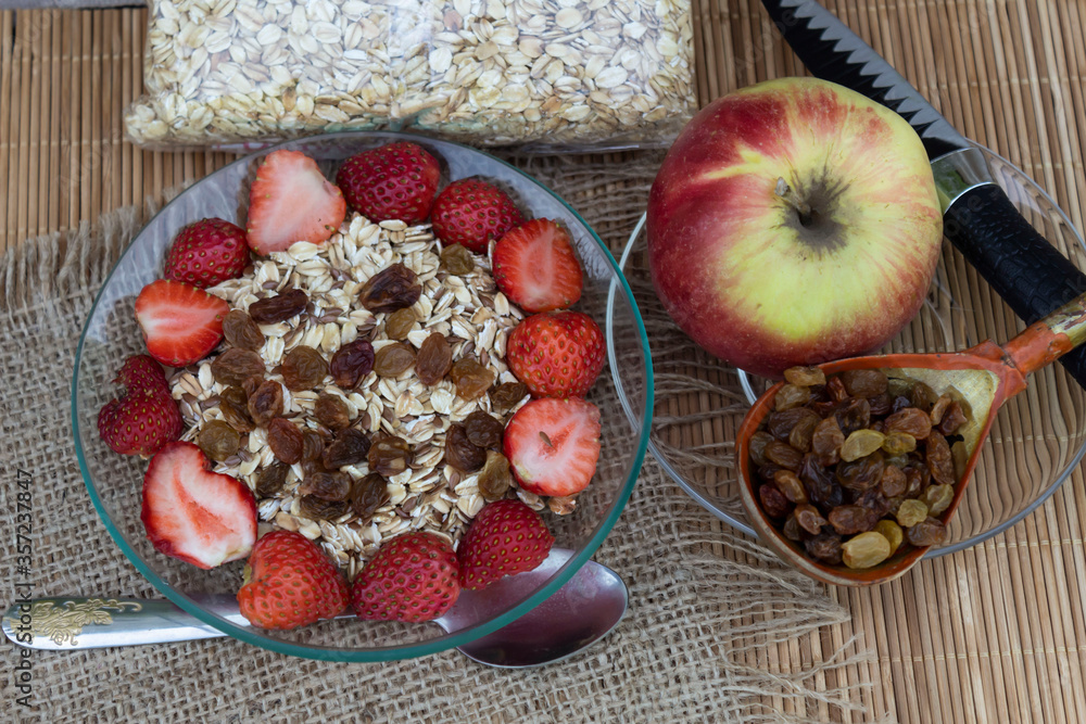 On the table is an apple, raisins in a wooden spoon, and oatmeal in a bag. Muesli with strawberries is cooked in a bowl. On the table is an apple, raisins in a wooden spoon, and oatmeal in a bag. Muesli with strawberries is cooked in a bowl.