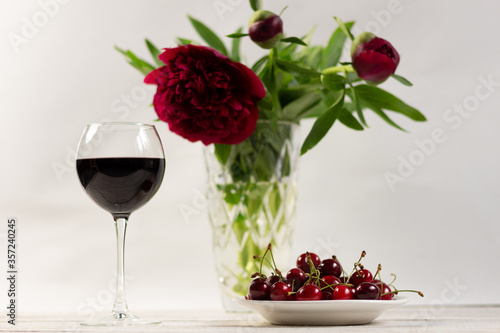 Red wine glass, ripe cherry berries in plate and peon flowers bouquet in vase on white wooden background