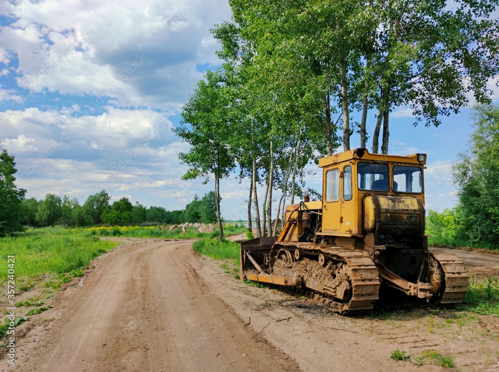 old yellow bulldozer stands on the side of a country road near trees against a blue sky with clouds on a sunny day old yellow bulldozer stands on the side of a country road near trees against a blue sky with clouds on a sunny day