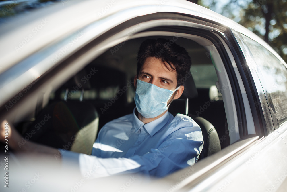 Young man sits behind the steering wheel in the car wearing sterile medical mask. Boy taxi driver works hard during coronavirus outbreak. Social distance, virus spread prevention and treat concept. Young man sits behind the steering wheel in the car wearing sterile medical mask. Boy taxi driver works hard during coronavirus outbreak. Social distance, virus spread prevention and treat concept.