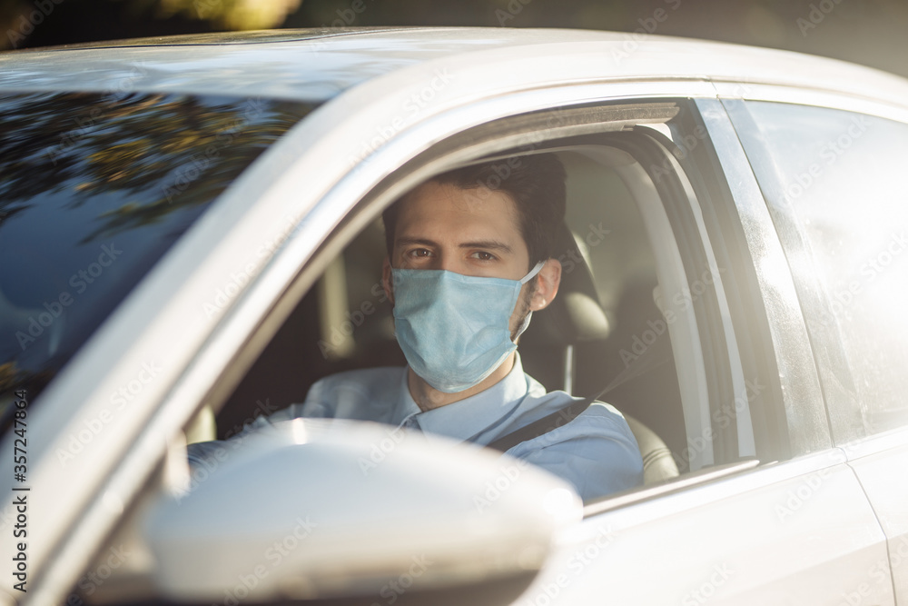 Fototapeta premium Closeup portrait of young man sits in the car wearing sterile medical mask. Boy taxi driver works hard during coronavirus outbreak. Social distance, virus spread prevention and treat concept.