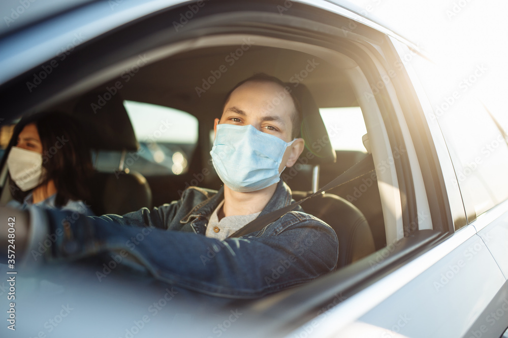 Young man drives a car with a passenger during coronavirus pandemic quaranteene. Taxi driver wears a mask sitiing behind the steering wheel. Social distance, epidemic, protection, health concept.