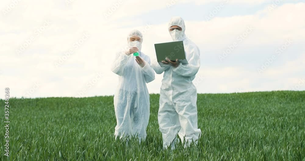 Caucasian female farmer researcher holding test tube with chemicals pesticides and male researcher with laptop computer in green field. Biologists genetics colleagues working outdoors in wheat margin. Caucasian female farmer researcher holding test tube with chemicals pesticides and male researcher with laptop computer in green field. Biologists genetics colleagues working outdoors in wheat margin.