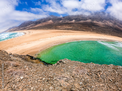 A view of a remote empty sandy beach, with volcanic mountains in the back; two bodies of sea emerge photo