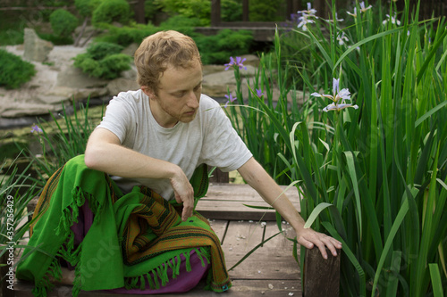 Young handsome man sits on a wooden bridge in the grass and reaches for a flower 