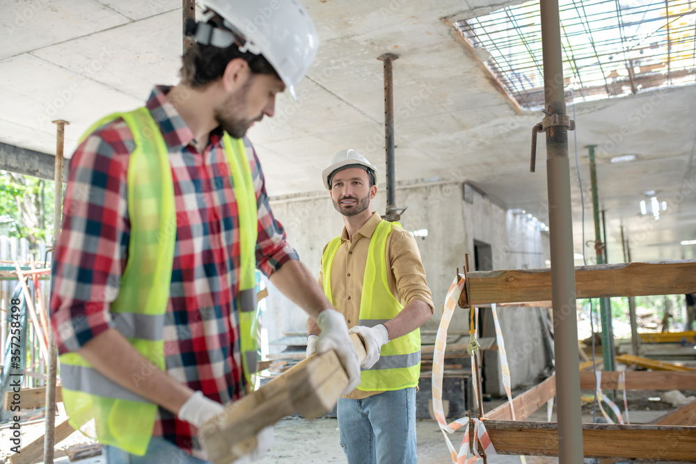Building workers in yellow vests and gloves carrying together wooden board Building workers in yellow vests and gloves carrying together wooden board
