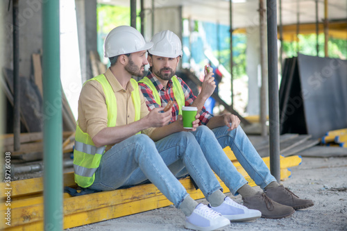 Building workers in yellow vests and helmets sitting on boards, having coffee with sandwiches, discussing something