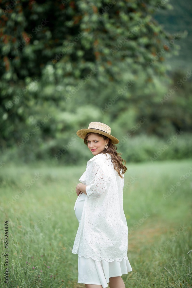 Portrait of a young beautiful pregnant woman. A pregnant woman in a straw hat walks in the park. Motherhood.