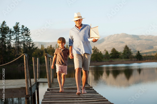 Smiling grandfather grandson toy sailboat holding hands walking along dock over lake
