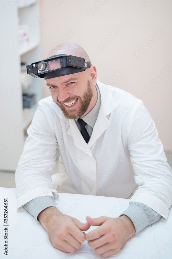 Male bearded doctor in a medical coat and with a magnifying head strap w lights sitting on the desk. Optometrist equipment. Medical office. Male bearded doctor in a medical coat and with a magnifying head strap w lights sitting on the desk. Optometrist equipment. Medical office.