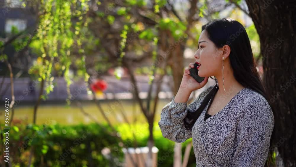 Chinese girl walking and talking on phone. China woman uses cellphone to make call whilst taking a walk in the park on a sunny day. Chinese girl walking and talking on phone. China woman uses cellphone to make call whilst taking a walk in the park on a sunny day.