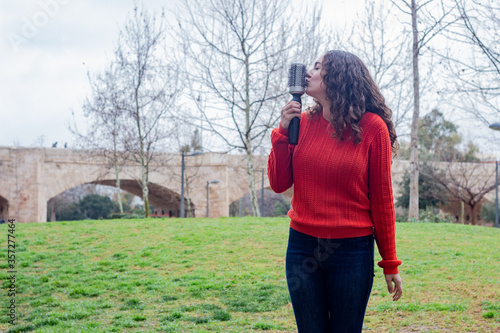 Portrait of caucasian young woman kissing rotating and styling brush, happy smiling, in the park, orange sweater and jeans, long curly hair. Place for your text in copy space.