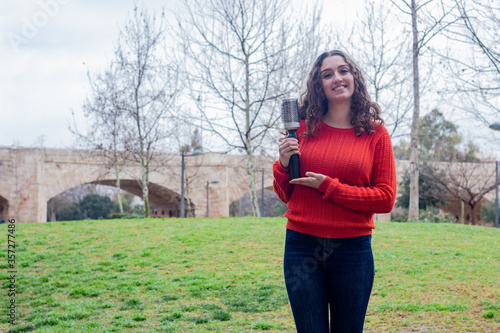 Portrait of attractive caucasian young woman model holding rotating and styling brush, in the park, orange sweater and jeans. Place for your text in copy space.