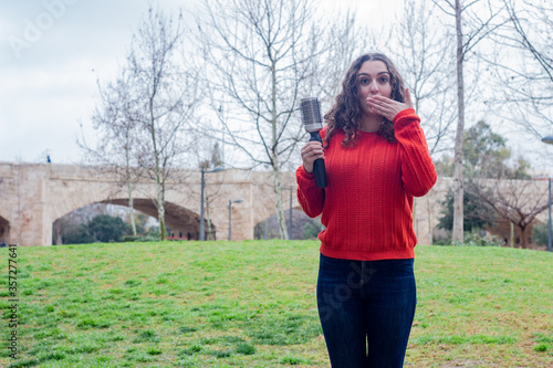 happy attractive caucasian young woman with rotating and styling brush . Covered mouth with hand smiling, in the park, orange sweater and jeans, long curly hair. Place for your text in copy space.