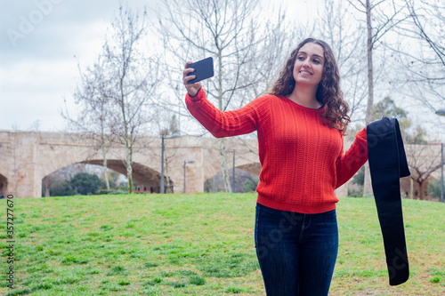 happy attractive caucasian young woman model holding electronic abdominal appliance, taking a selfie, in the park, orange sweater and jeans, long curly hair. Place for your text in copy space.