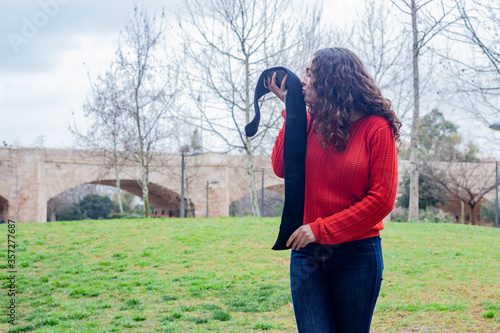 Portrait of caucasian young woman model kissing electronic abdominal appliance, happy smiling, in the park, orange sweater and jeans, long curly hair. Place for your text in copy space.