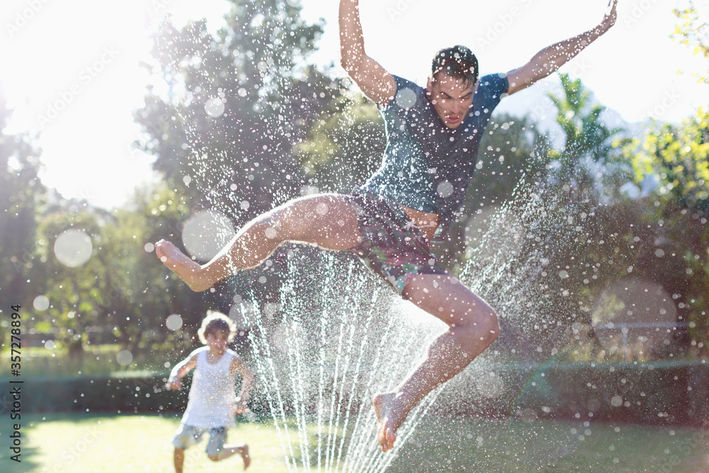 Father and son playing in sprinkler in backyard - obrazy, fototapety, plakaty 