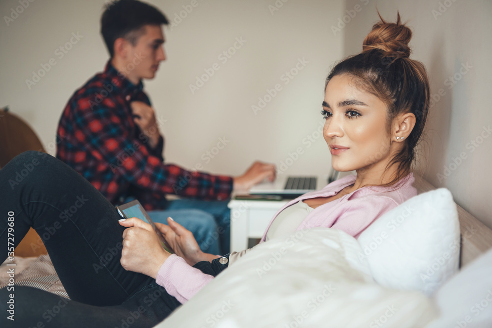 Fototapeta premium Young caucasian couple sitting in the bedroom and using modern devices working and spending time together