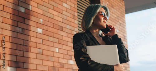 Caucasian business director talking on phone outside while holding a laptop in a brick wall