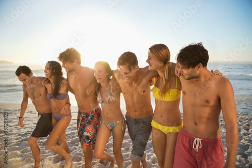 Friends in bikinis and swim trunks hugging and walking on beach
