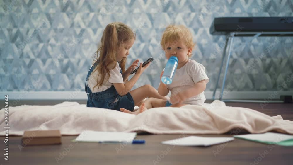 funny little boy with bottle of water sits near elder sister playing with modern mobile phone on blanket on floor in spacious room funny little boy with bottle of water sits near elder sister playing with modern mobile phone on blanket on floor in spacious room