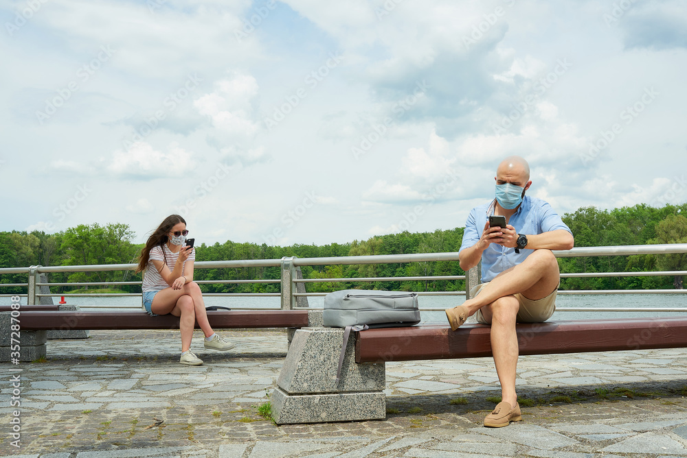 A bald man in a medical face mask is sitting on a bench keeping a social distance with a woman sitting on another bench on the promenade. A guy and a girl are using smartphones in social distancing.