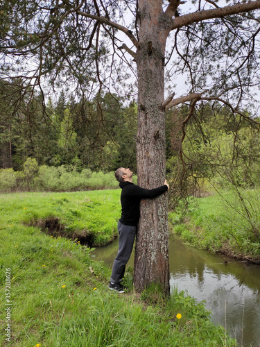 A young man is resting in the forest near a stream or lake. The man embraces a old tree on a forest glade.