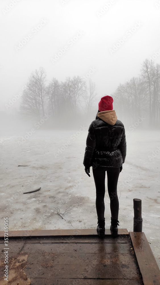 A young woman stands alone on a wooden bridge by the lake. Early cloudy foggy day. To be alone with yourself. Tight fog.