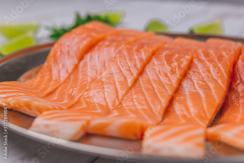 Raw vibrant salmon fillet in a serving plate with herbs, creamy sauce made from butter, garlic, and vegetables. Salmon with wedge lime on the side on a white wooden table.
