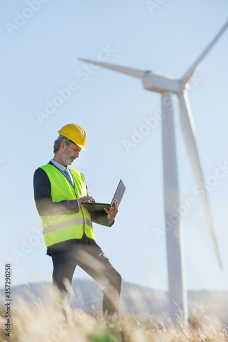 Worker using laptop by wind turbine in rural landscape photo