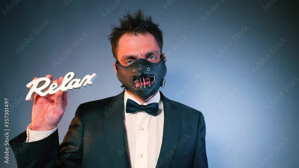 The dominance of behavior. Therapy of the psyche. A masked man in a black suit shows the word relax. In the other hand holds a sex toy. BDSM session preparation concept. The dominance of behavior. Therapy of the psyche. A masked man in a black suit shows the word relax. In the other hand holds a sex toy. BDSM session preparation concept.