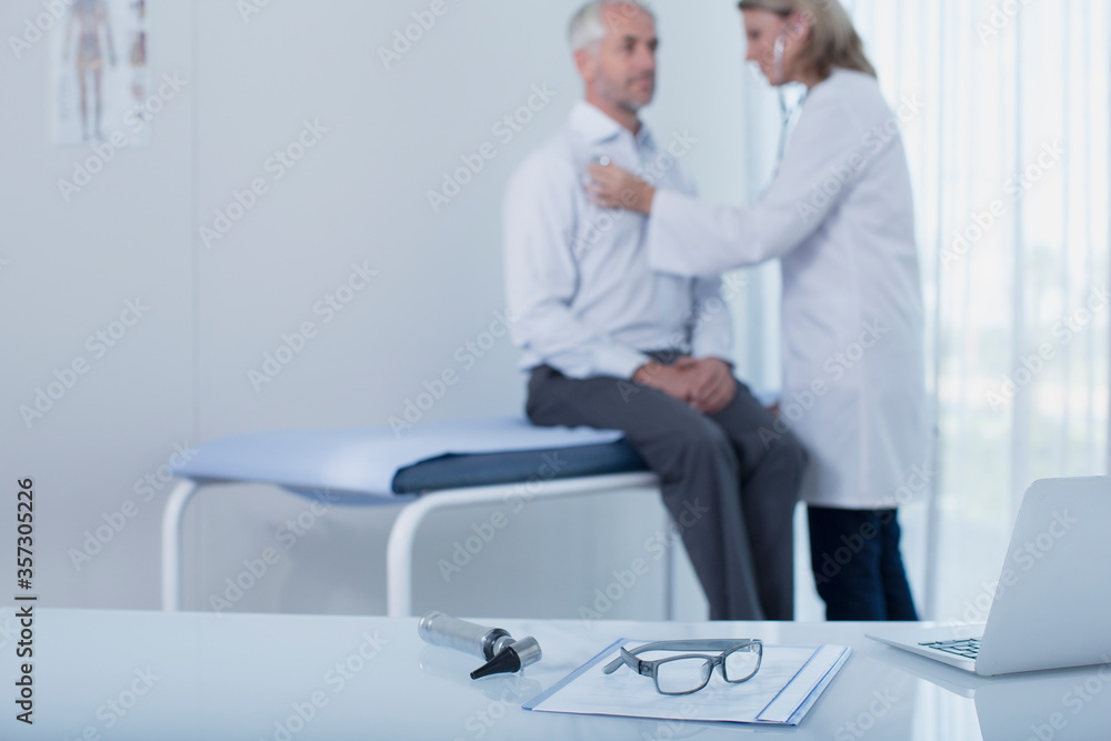 Female doctor examining patient in office, laptop, file, glasses otoscope on desk in foreground