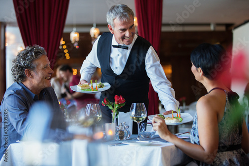 Smiling waiter serving fancy dishes to mature couple sitting at table in restaurant
