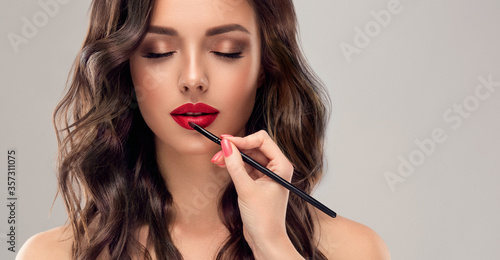 Makeup artist applies red lipstick . Beautiful woman face. Hand of visagiste, painting cosmetics of young beauty model girl . Make up in process