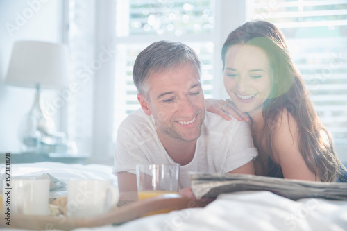 Smiling couple reading newspaper in sunny bedroom