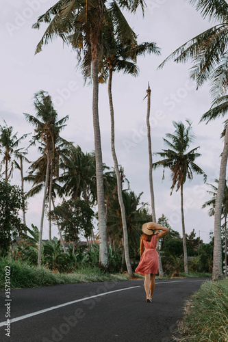 A young beautiful woman in a dress and hat walks along the road with palm trees © Denis Chernousov